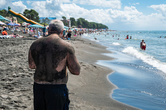 Man covered in black magnetic sand stands on the sunny beach in Ureki, Georgia, during a summer vacation by the Black Sea.