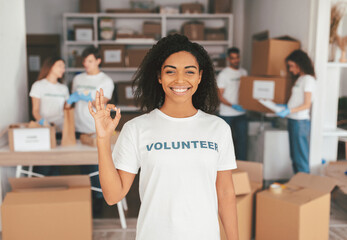 A group of volunteers are actively preparing donation boxes in a community center. One smiling volunteer in the foreground holds up a gesture of approval. The atmosphere is positive and collaborative.