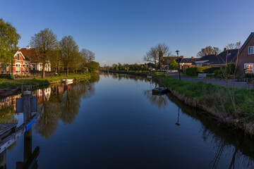 Obraz premium A wide view of the village of Bedum in the Netherlands, where brick houses, grassy banks, and trees are reflected in the calm canal under a clear sky