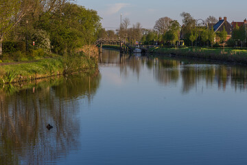 A wide view of a calm canal lined with lush green vegetation in Bedum. A wooden bridge and traditional Dutch houses are visible in the background. A peaceful morning scene