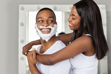 A young man with shaving cream on his face smiles at a young woman hugging him from behind. They are in a bright bathroom decorated with a large mirror, enjoying a fun moment together.