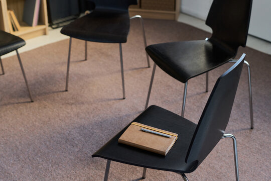 Empty chairs arranged in circle with notebook and pen resting on seat, representing group therapy session setup in neutral indoor space, no people in scene