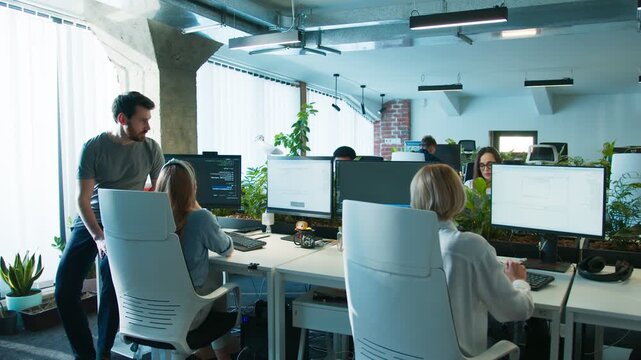 Male standing beside coworker. Team coding at desks in open workspace. Several workers focusing on monitors while developing software. Office area filled with plants and computers.