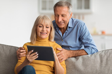 A happy older couple sits closely on a sofa, smiling as they look at a tablet together. They are in a bright living room filled with warmth and comfort.