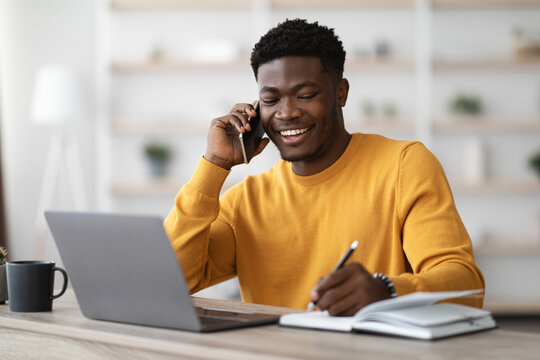 Happy african american young man freelancer or businessman working from home, sitting at table in front of modern laptop, having phone conversation, taking notes and smiling, copy space - Powered by Adobe