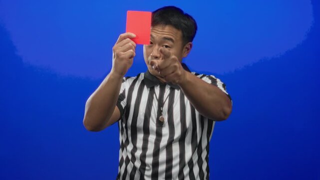 Young male chinese referee holds red card, blows whistle and points finger in blue studio; authority.