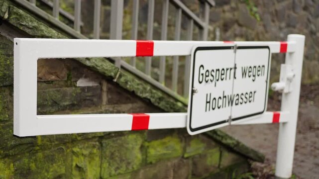 flood closure sign on white barrier showing german text 'gesperrt wegen hochwasser', red reflective stripes on horizontal rails, metal railing beside mossy stone wall, closed stairway and blocked