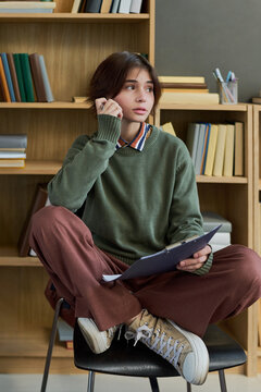Young woman sitting cross legged on chair holding clipboard, listening attentively during group therapy session, bookshelf with books in background