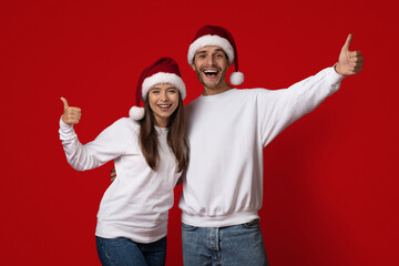 A happy couple wears matching Christmas hats while posing against a bright red background. They...