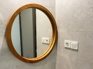 Round wooden-framed mirror hanging on a light gray tiled bathroom wall next to white electrical outlets. Modern interior design, home decor, and bathroom renovation.