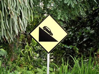 Black and yellow diamond warning sign indicating a steep slope or hill descent, set outdoors in a lush, green, tropical forest environment. Road safety concept.