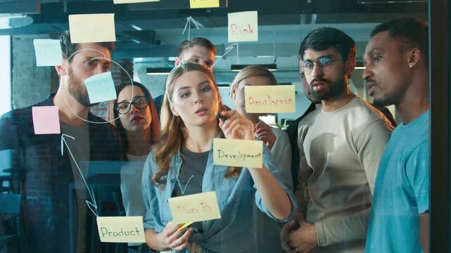 Female drawing arrows on glass board. Lead woman marking connections between steps like risk and analysis. Coworkers watching closely while discussing workflow. Bright planning room.