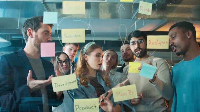 Team placing sticky notes on glass. Group focusing on workflow steps. Writing tasks while planning project stages. Office with collaborative teamwork atmosphere. Working on new ideas.