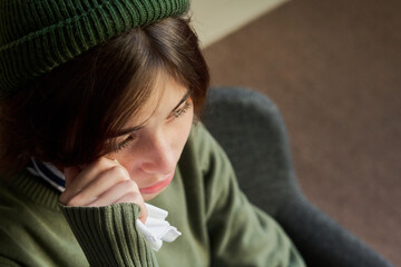 Portrait of young woman sitting in group therapy session holding tissue to face appearing emotional, wearing knit hat and sweater, looking downward thoughtfully