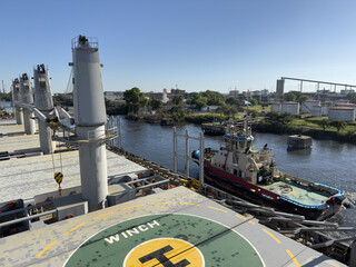 Tugboat assisting a large cargo ship in a river port, view from the deck with winch platform, maritime towing operations and harbor logistics.
