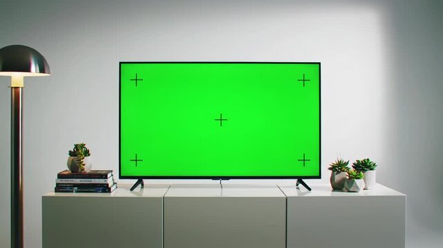 Static video of modern flat-screen tv with vivid green screen on polished white console, minimalist studio with lamp, books, plants. Versatile content placeholder concept