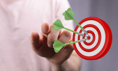 Close-up image of a person's hand with a dartboard, three darts perfectly hitting the target,...