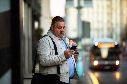Adult man focused on smartphone while eating at city bus stop