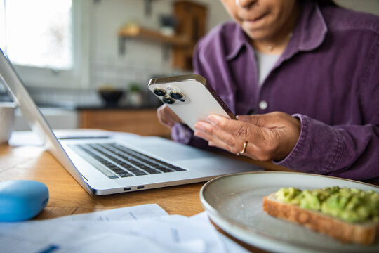 Adult woman focused on smartphone and laptop in home kitchen