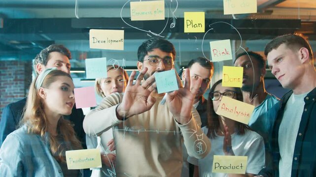 Coworker placing idea note on glass board while team observing. Men and women reviewing project structure closely. Group shaping workflow during detailed planning session inside office.