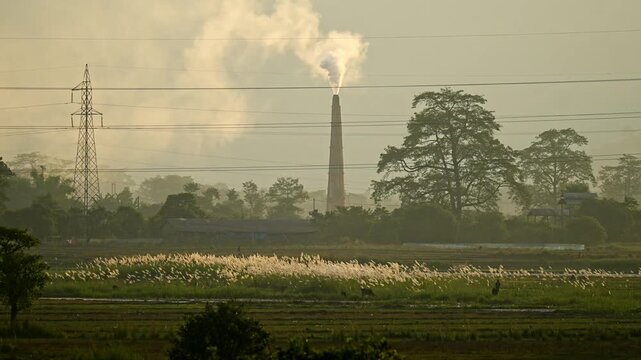 A brick kiln chimney releases smoke into the evening sky as it rises over fields and distant hills, highlighting the contrast between rural landscapes and industrial pollution
