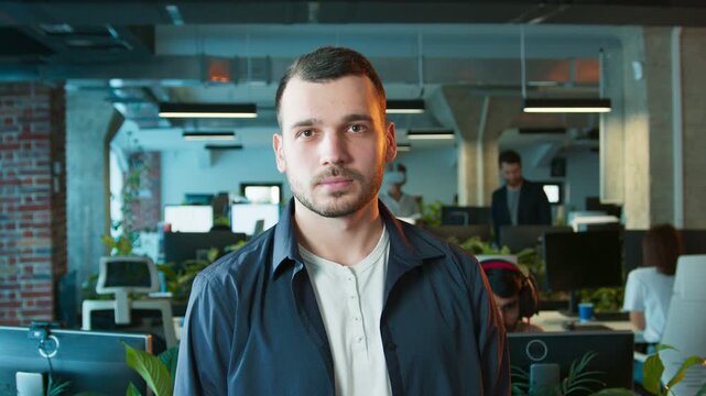Attractive male standing still while facing camera. Man posing confidently with neutral expression. Professional worker captured in office environment featuring monitors and plants in background.