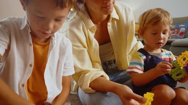 Young smiling mother and her two sons playing with a colorful constructor puzzle on the floor in the living room, enjoying a creative and educational activity together on a family weekend