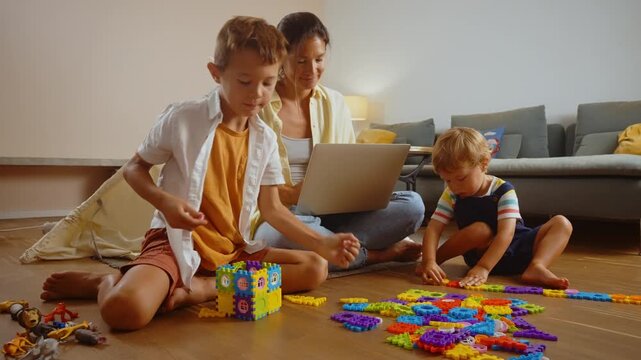 Young smiling mother and her two sons playing with a colorful constructor puzzle on the floor in the living room, enjoying a creative and educational activity together on a family weekend