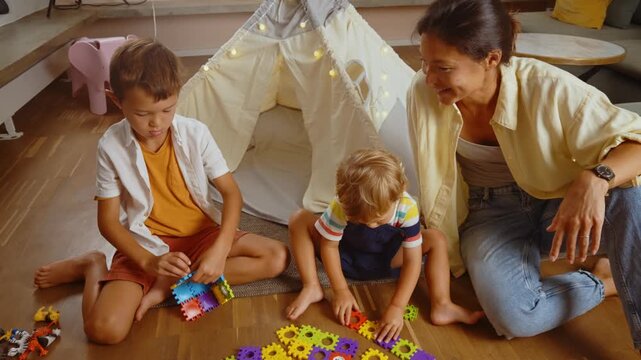 Young smiling mother and her two sons playing with a colorful constructor puzzle on the floor in the living room, enjoying a creative and educational activity together on a family weekend