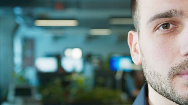 Camera close-up portrait of serious Caucasian male. Partial face showing eye and beard while looking at camera. Detailed facial texture in focus. Office environment with blurred background.