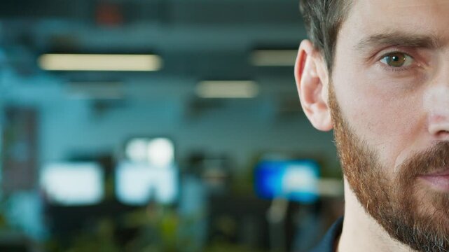 Close view of male professional inside office. Man looking directly ahead with serious expression. Focus placed on half of his face while blurred background showing active workspace behind him.