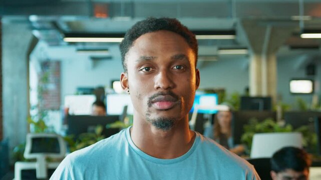 Focused African American male looking directly at camera. Man keeping neutral expression while standing in office. Coworkers working behind him during active day inside bright workspace.