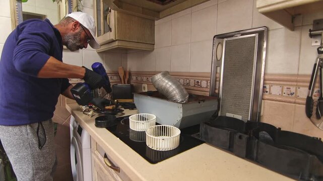 Man using a cordless drill to tighten screws on a disassembled range hood motor. Kitchen appliance service, DIY repair, and home maintenance work.