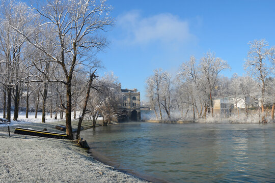 A scenic winter view of the Kupa River at the Slava Raskaj swimming area in Ozalj, featuring frost-covered trees and the historic Munjara hydroelectric power plant in the distance under a bright blue 
