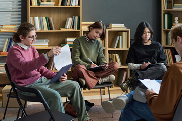 Group of multiethnic teenagers sitting in circle participating in group therapy session, holding clipboards and writing notes, focused on discussion, bookshelves in background © Mediaphotos