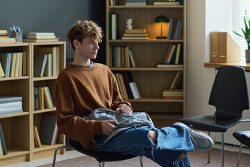 Teenage boy sitting alone in group therapy room holding backpack on lap looking to side appearing thoughtful surrounded by bookshelves and empty chairs