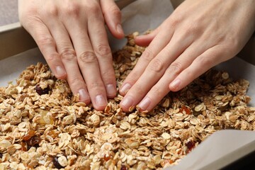 Making granola. Woman putting oats flakes with dried fruits onto tray at grey table, closeup