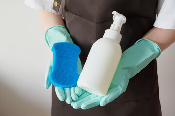 Woman wearing an apron and rubber gloves holding blue sponge and white soap dispenser.