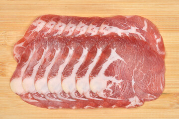 An overhead shot of approximately nine thin slices of dry-cured smoked ham or Coppa, showing clear marbling, arranged on a light wood cutting board