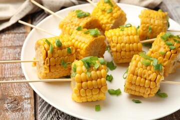 Skewers with pieces of boiled corncobs and green onion on wooden table, closeup
