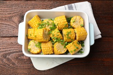 Pieces of boiled corncobs with green onion in baking dish on wooden table, top view