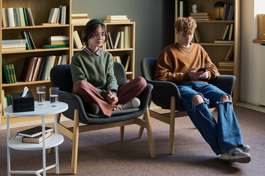 Young woman sitting cross legged on chair looking forward while young man sitting beside her using smartphone during group therapy session in room - Powered by Adobe