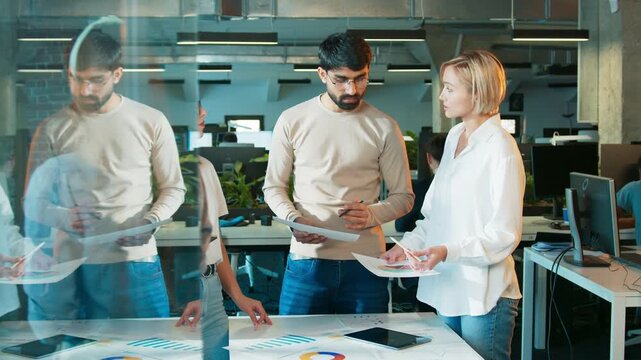 Three colleagues standing near table. Man reviewing printed report. Two women studying charts and tablet. Team planning next steps in office room. Papers showing data on desk surface.