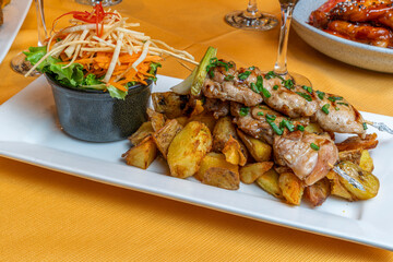 Grilled chicken skewers with roasted potatoes and fresh salad served on a white plate in a restaurant setting with wine glasses on a yellow tablecloth.