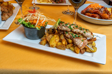 Grilled chicken skewers with roasted potatoes and fresh salad served on a white plate in a restaurant setting with wine glasses on a yellow tablecloth.