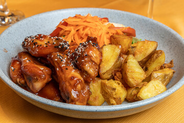 Sesame glazed chicken wings with roasted potatoes and fresh salad served in a ceramic bowl on a yellow tablecloth in a restaurant setting.