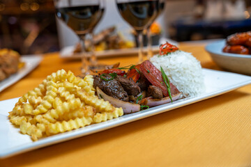 Peruvian lomo saltado with sautéed beef, onions and tomatoes served with white rice and crispy crinkle fries on a bright tablecloth in a restaurant setting.