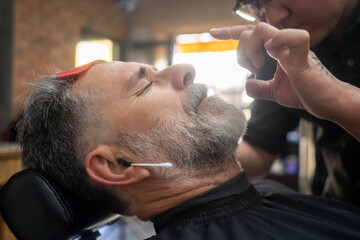 Barber grooming eyebrows of mature man with razor in barber shop