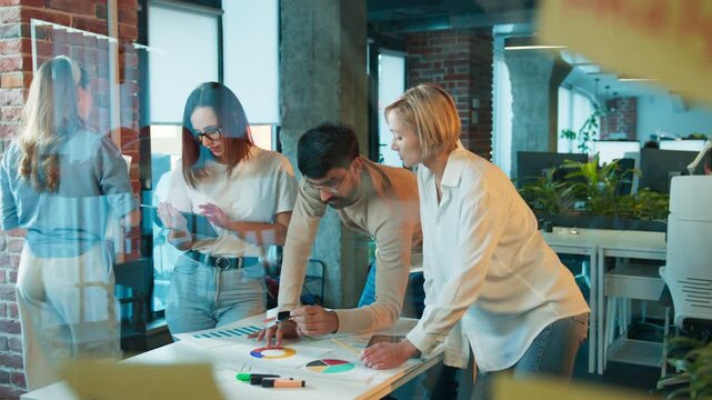 Three colleagues leaning over table. Man reviewing charts with marker. Two women checking notes and tablet. Team planning project in office room. Data sheets covering desk surface.