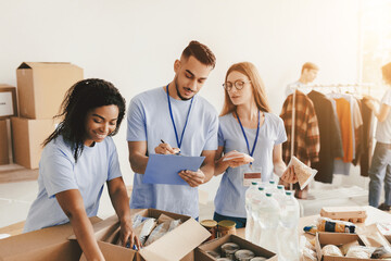 Three volunteers work together in a community service center, sorting food donations and preparing boxes. They display teamwork while coordinating items and writing notes for the distribution.